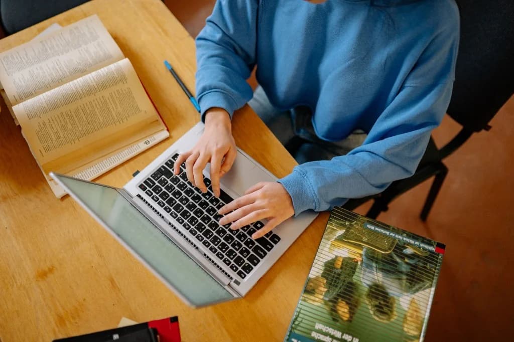 Student typing on a laptop at a desk with textbooks and study materials