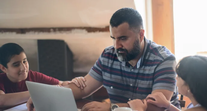 Parent at a laptop with two children nearby, focused on test prep resources together